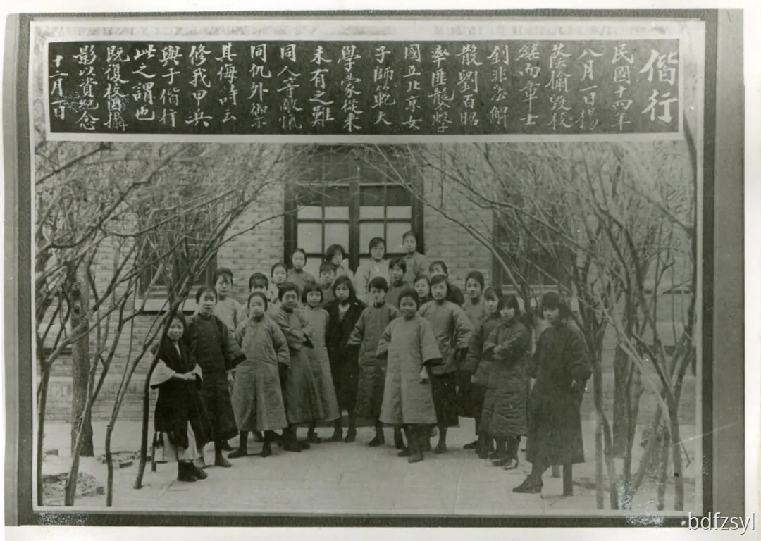 A group of young Chinese women or girls in long coats pose for a historical black and white photograph outdoors in front of a building, framed by bare trees and a large banner with Chinese calligraphy. (Captioned by AI)