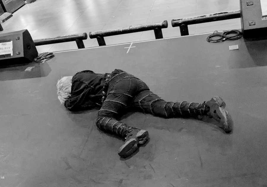 A person with bleached hair and black clothes lies face down on a dark floor next to some stage equipment. (Captioned by AI)