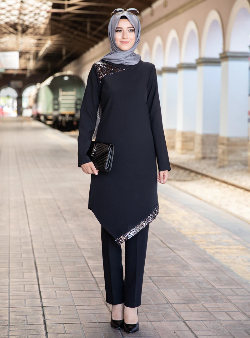 在火车站台背景下，一名穿着深蓝色上衣、黑色长裤和灰色希贾布的女性正对着镜头微笑，手中拿着一个黑色的手拿包。 (由 AI 生成标题)